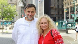 A man in a white embroidered linen shirt stands next to a woman with curly blonde hair in a red vyshyvanka dress decorated with white cross-stitch patterns. Both smile at the camera on a sunny plaza in downtown Manhattan, with green trees, a subway entrance and old buildings behind them. People gather in downtown Manhattan for a Ukrainian rally, waving blue and yellow flags and showing solidarity and support for Ukraine in the streets of New York City. Photo: Olek Buzunov (@buzunov.photoart)